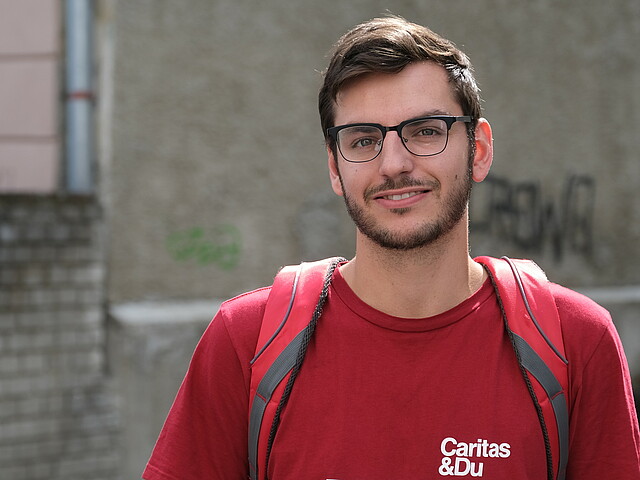 Portrait eines jungen Mannes mit Brille, rotem Caritas T-Shirt und Rucksack.