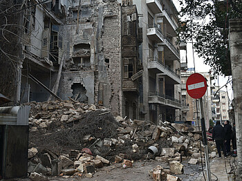 Eine Gruppe Personen steht neben einem stark zerstörten Gebäude in der syrischen Stadt Aleppo.