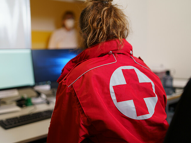 Eine Frau sitzt an einem Empfangstisch mit Bildschirm und trägt eine Einsatzjacke des Roten Kreuzes.