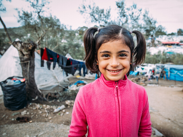Portrait eines Mädchens mit pinkem Fleece-Pullover und langen Zöpfen, das mitten in in einem Zeltlager für Flüchtlinge steht.