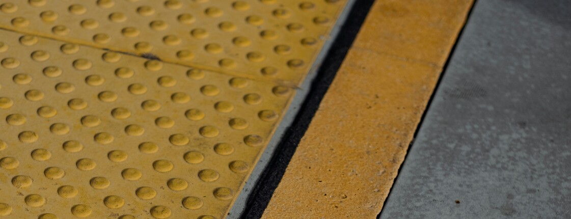 Barrierefreiheitserklärung Nahaufnahme eines Randsteins, der mit gelber Farbe und einer genoppten Oberfläche für Menschen mit Beeinträchtigungen markiert wurde.