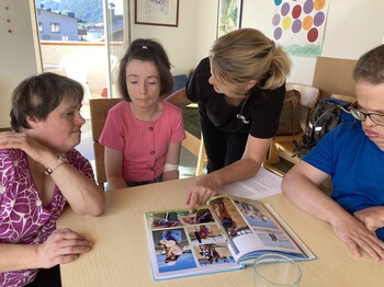Drei Menschen mit Behinderungen sitzen an einem Tisch, auf dem ein aufgeschlagenes Fotoalbum liegt. Eine Caritas Mitarbeiterin zeigt auf eines der Fotos.