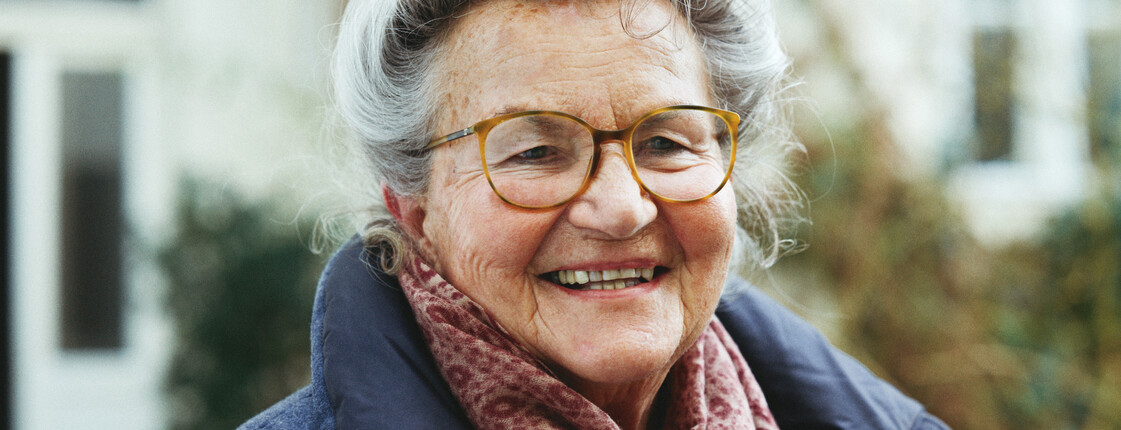 Portrait einer Seniorin mit Brille, die einen roten Schal und einen blauen Wintermantel trägt.