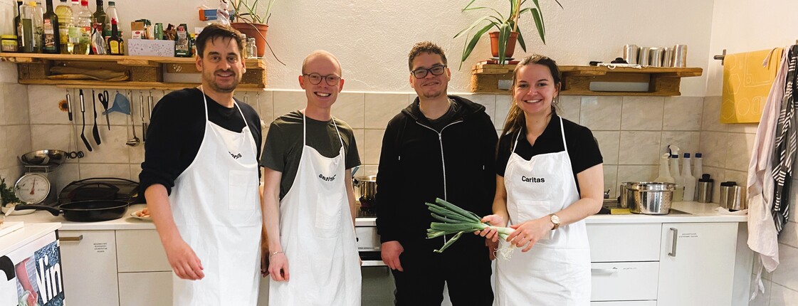 Vier Personen mit weißen Kochschürzen stehen nebeneinander in einer Küche.