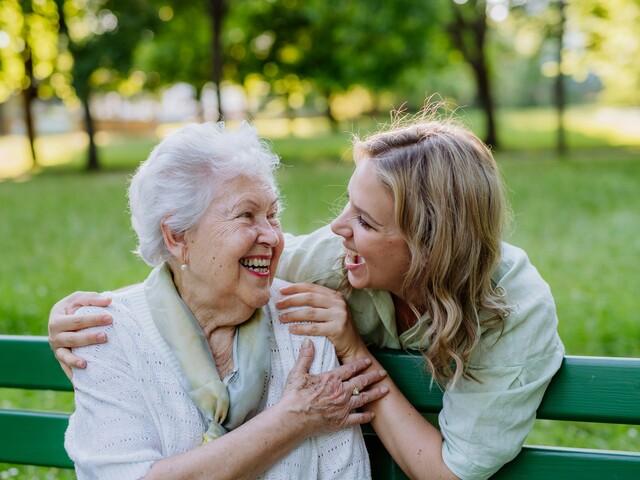 In einem Park sitzt eine Seniorin auf einer grünen Bank. Hinter ihr steht eine junge Frau, die ihre Hände auf die Schultern der Seniorin gelegt hat. Beide schauen sich lachend an.