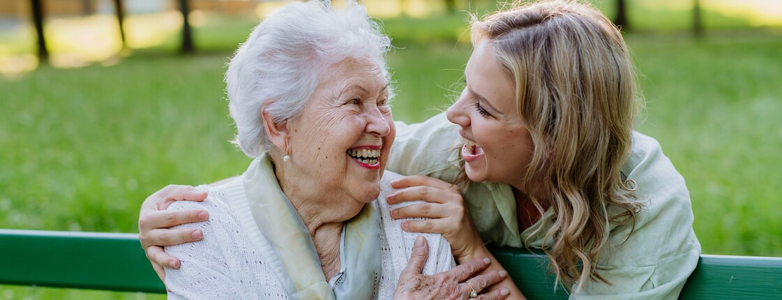 In einem Park sitzt eine Seniorin auf einer grünen Bank. Hinter ihr steht eine junge Frau, die ihre Hände auf die Schultern der Seniorin gelegt hat. Beide schauen sich lachend an.