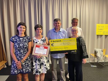 Fünf Personen stehen bei der Spendenübergabe zu einem Gruppenfoto zusammen und halten einen Spendenscheck hoch.