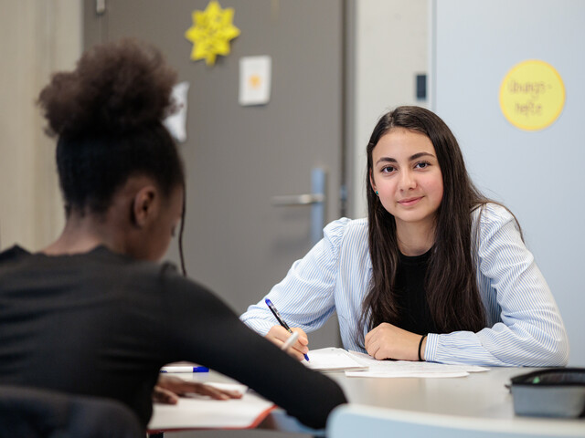 Portrait einer Schülerin, die mit einer anderen Schülerin an einem Tisch Hausaufgaben macht.
