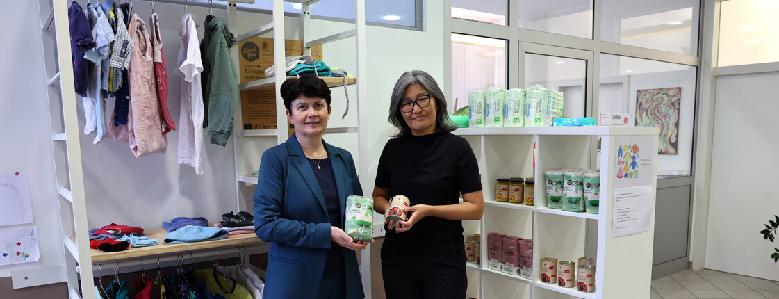 Zwei Frauen stehen in einem großen, hellen raum vor zwei weißen Regalen mit Sachspenden.