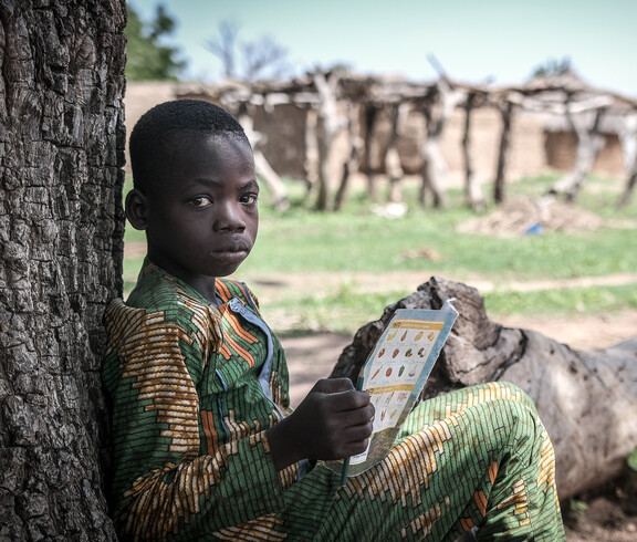 Portrait eines afrikanischen Jungen, der an einem Baum lehnend sitzt, und in einem Heft liest.