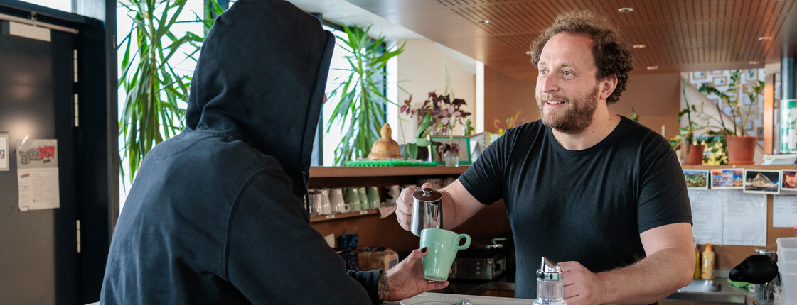 Ein Caritas freundlicher Mitarbeiter in einem Tageszentrum schenkt einem Klienten Kaffee nach.