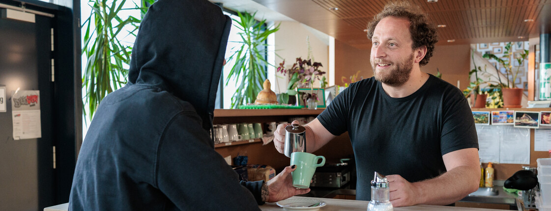 Ein Caritas freundlicher Mitarbeiter in einem Tageszentrum schenkt einem Klienten Kaffee nach.