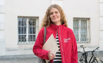 Portrait einer Caritas Mitarbeiterin, die eine rote Caritas Jacke und ein Holzklemmbrett unter dem Arm trägt, vor einem Gebäude.