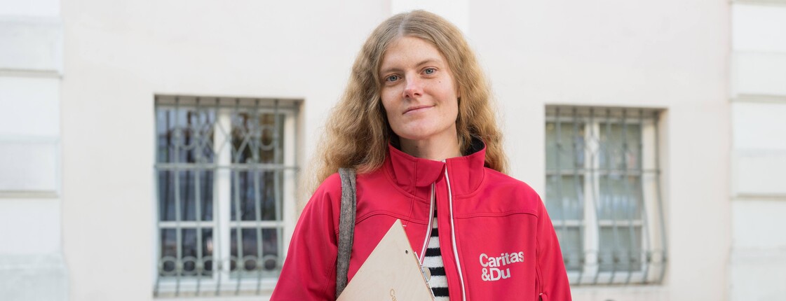 Portrait einer Caritas-Mitarbeiterin mit roter Jacke, einem Stoffbeutel und einem Klemmbrett mit Caritas-Logo. Im Hintergrund ein Wohnhaus. 