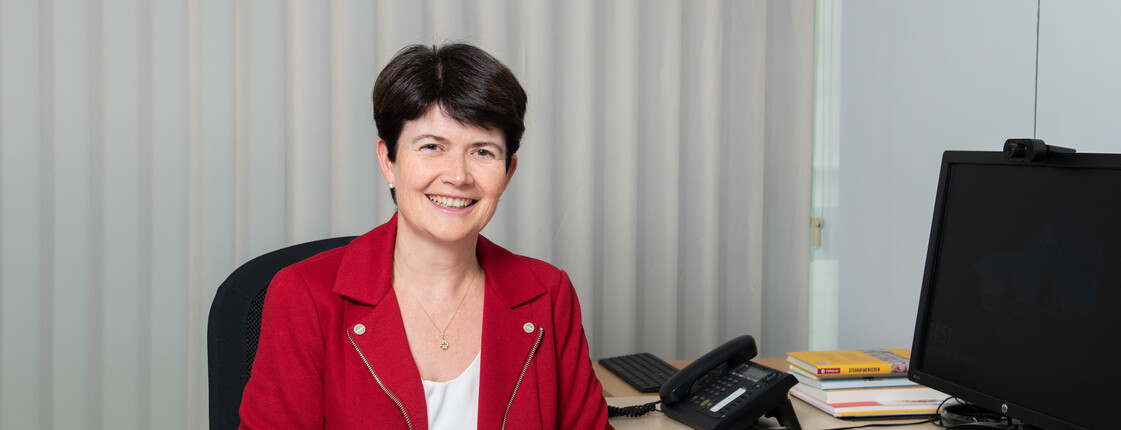 Portrait einer am Schreibtisch sitzenden Frau, die eine rote Jacke trägt.