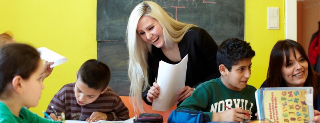 Lerncafé Schulkinder sitzen an einem Tisch und machen Aufgabe, daneben steht eine junge Frau mit Zetteln in der Hand.