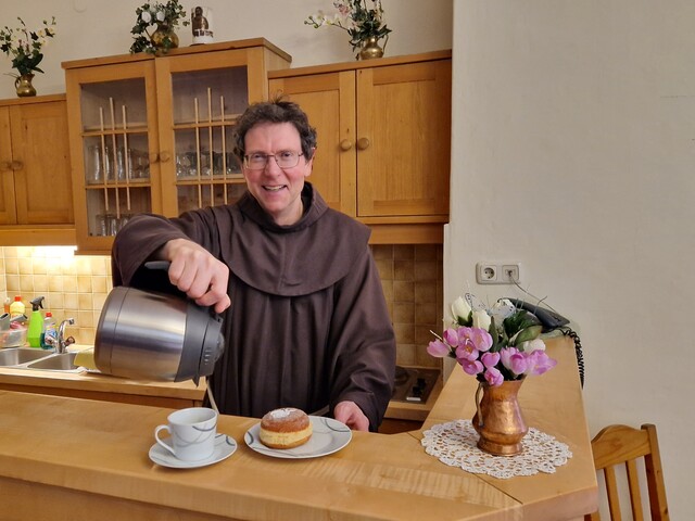Ein Pater mit brauner Kutte steht hinter einem Holztressen, auf dem ein Teller mit einem Krapfen steht, und schenkt aus einer Thermoskanne Wasser in eine Tasse.