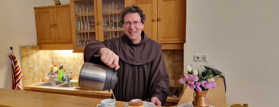 Teestube Lienz  Ein Pater mit brauner Kutte steht hinter einem Holztressen, auf dem ein Teller mit einem Krapfen steht, und schenkt aus einer Thermoskanne Wasser in eine Tasse.