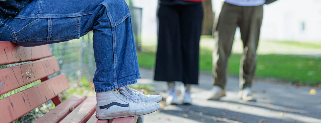 Eine Frau, die eine blaue Jeans und einen Sweatshirt sowie weiße Sneaker trägt, sitzt auf der Rücklehne einer roten Parkbank. Im Hintergrund nähern sich ihr zwei Caritas Mitarbeiter*innen.