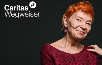 Das Porträt einer Frau ist zu sehen auf schwarzem Hintergrund. Es ist eine ältere Dame die lächelt, sie hat einen rothaarigen Kurzhaarschnitt. Links oben das Logo vom Caritas Wegweiser.