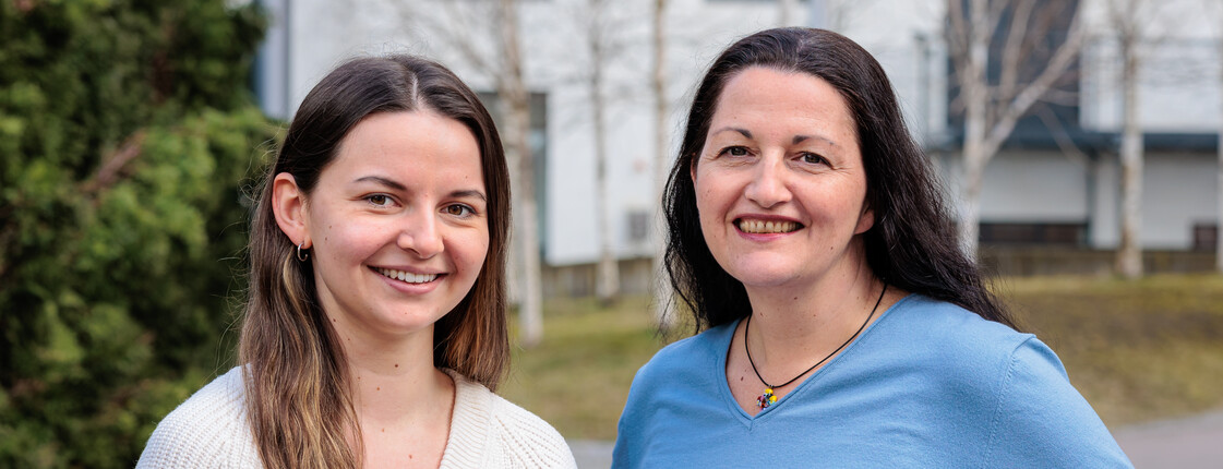 Portrait zweier lächelnder Frauen mittleren Alters, die in einer Fußgängerzone zu einem Portrait zusammen stehen.