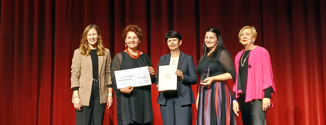 Fünf festliche gekleidete Frauen stehen auf einer Bühne vor einem roten Vorhang und präsentieren Zertifikate.