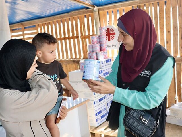 Eine Mitarbeiterin der Caritas Jerusalem zeigt einer Frau mit kleinem Kind eine Dose Babynahrung. Im Hintergrund sind Hilfspakete und das Caritas-Logo zu sehen.