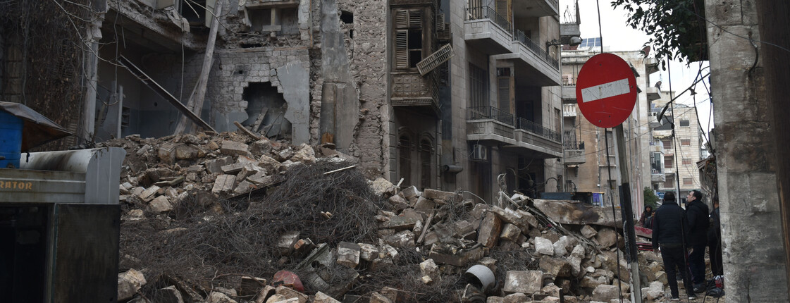 Eine Gruppe Personen steht neben einem stark zerstörten Gebäude in der syrischen Stadt Aleppo.
