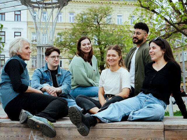 Sechs junge Erwachsene und eine ältere Person sitzen lachend im Park und blicken sich an