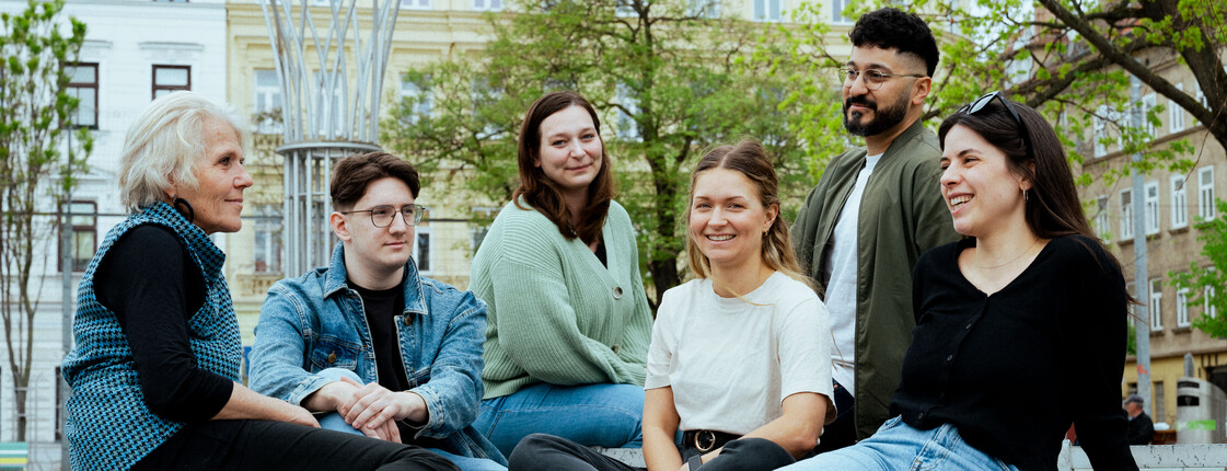Sechs junge Erwachsene und eine ältere Person sitzen lachend im Park und blicken sich an