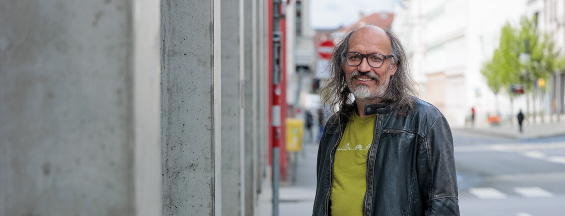 Portrait eines Caritas Mitarbeiters, der neben einer Gebäudeaußenfassade aus Beton steht und die Hände in den Hosentaschen hat.