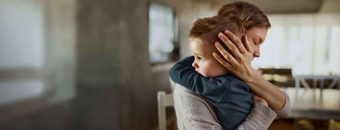 Spendenmöglichkeiten Eine Frau, die in einem Wohnzimmer auf einem Stuhl sitzt und ihre Augen geschlossen hat, hält ein Kind in den Armen. Das Kind schaut aus dem Fenster.