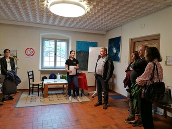 In einem Raum stehen einige Personen um eine Caritas-Mitarbeiterin, welche einen Vortrag hält.
