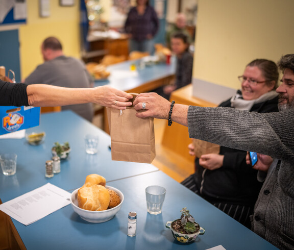 Eine Frau reicht einem Mann eine Papiertüte mit Essen über einen Tisch.