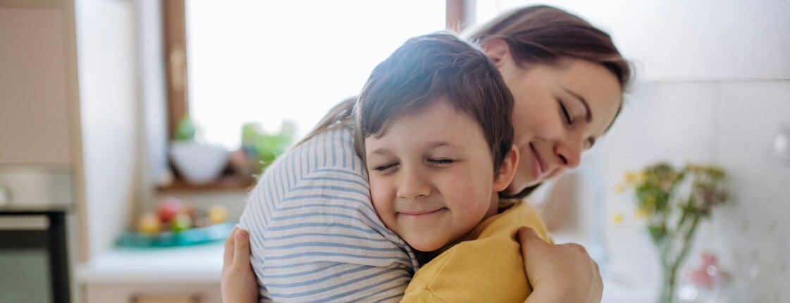Eine Mutter umarmt ihren Sohn, der ein gelbes T-Shirt trägt, in einer von natürlichem Licht durchfluteten Küche.