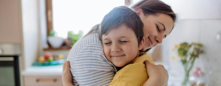 Eine Mutter umarmt ihren Sohn, der ein gelbes T-Shirt trägt, in einer von natürlichem Licht durchfluteten Küche.
