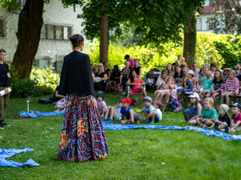 In einem Garten steht eine Frau vor zuschaunenden Kindern.