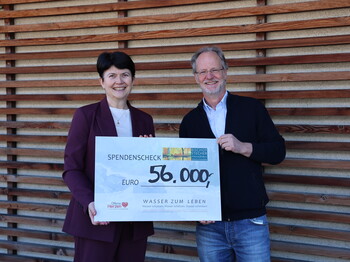 Eine Frau und ein Mann stehen vor einer holzverkleideten Wand nebeneinander und präsentieren einen Spendenscheck über 65.000 Euro.