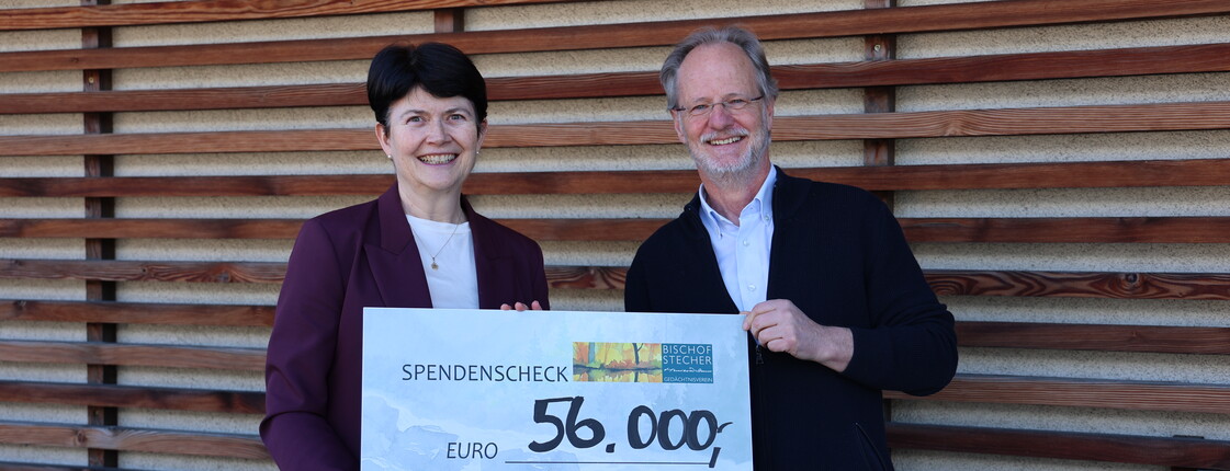 Eine Frau und ein Mann stehen vor einer holzverkleideten Wand nebeneinander und präsentieren einen Spendenscheck über 65.000 Euro.