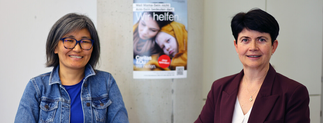 Zwei Frauen stehen an einem Stehtisch. Im Hintergrund ein Plakat der Caritas Inlandskampagne.
