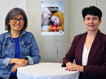 Zwei Frauen stehen an einem Stehtisch. Im Hintergrund ein Plakat der Caritas Inlandskampagne.