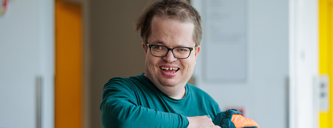 Ein lachender Mann mit Behinderung und Brille steht in einer Tagesstätte für Menschen mit Behinderung am Empfangstresen.
