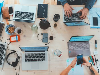 Obersicht eines modernen Schreibtisches aus hellem Holz, an dem fünf Personen mit Laptops und weiteren Bürogeräten arbeiten.