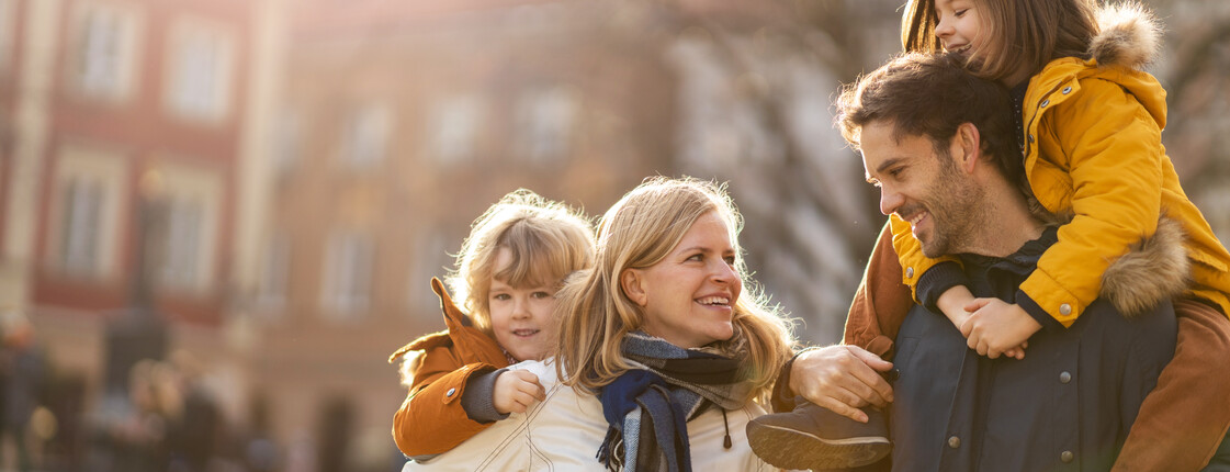 Familien- und Lebensberatung Eine gut gelaunte Familie in Winterkleidung geht bei Sonnenschein durch die Stadt. Vater und Mutter tragen jeweils ein Kind auf den Rücken.