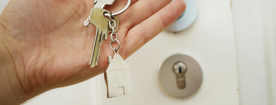 Housing First Vor einer weißen Wohnungstür hält eine Hand einen Schlüsselbund mit zwei Schlüsseln und einer Schlüsselanhänger, der ein Haus darstellt, hoch.