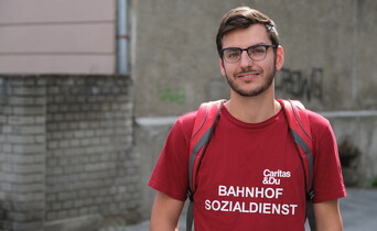 Straßenportrait eines Mannes mit Brille, der ein rotes Caritas T-Shirt und einen roten Rucksack trägt.