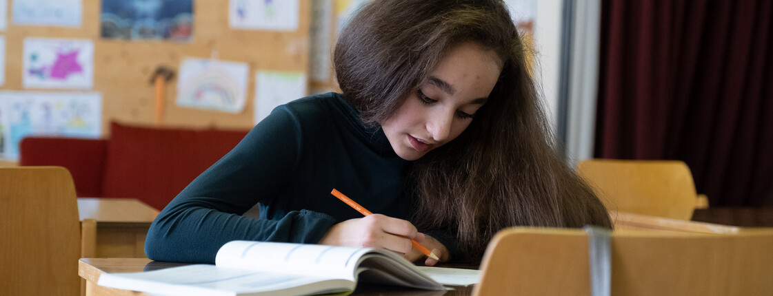 Ein Mädchen sitzt an einem Tisch und lehnt konzentriert über einem Schulaufgabenheft.