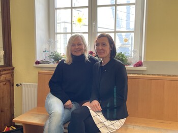 Zwei Frauen sitzen vor einem großen Fenster nebeneinander auf einer Holzbank