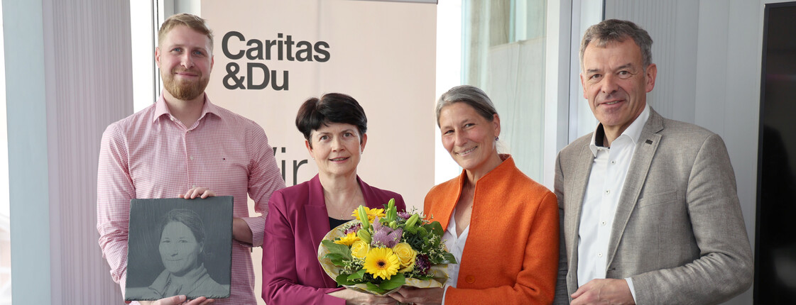 Vier Personen stehen vor einem Caritas Roll-Up zu einem Pressefoto zusammen. Die beiden Frauen in der Bildmitte halten einen Blumenstrauß.