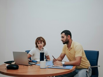 Eine Frau und ein Mann sitzen an einem runden Holztisch, auf dem Schreibunterlagen liegen, und schauen in einen Laptop.